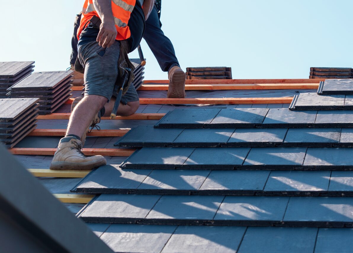 Man wearing an orange vest and safety vest working on a roof, holding a hammer in his hand
