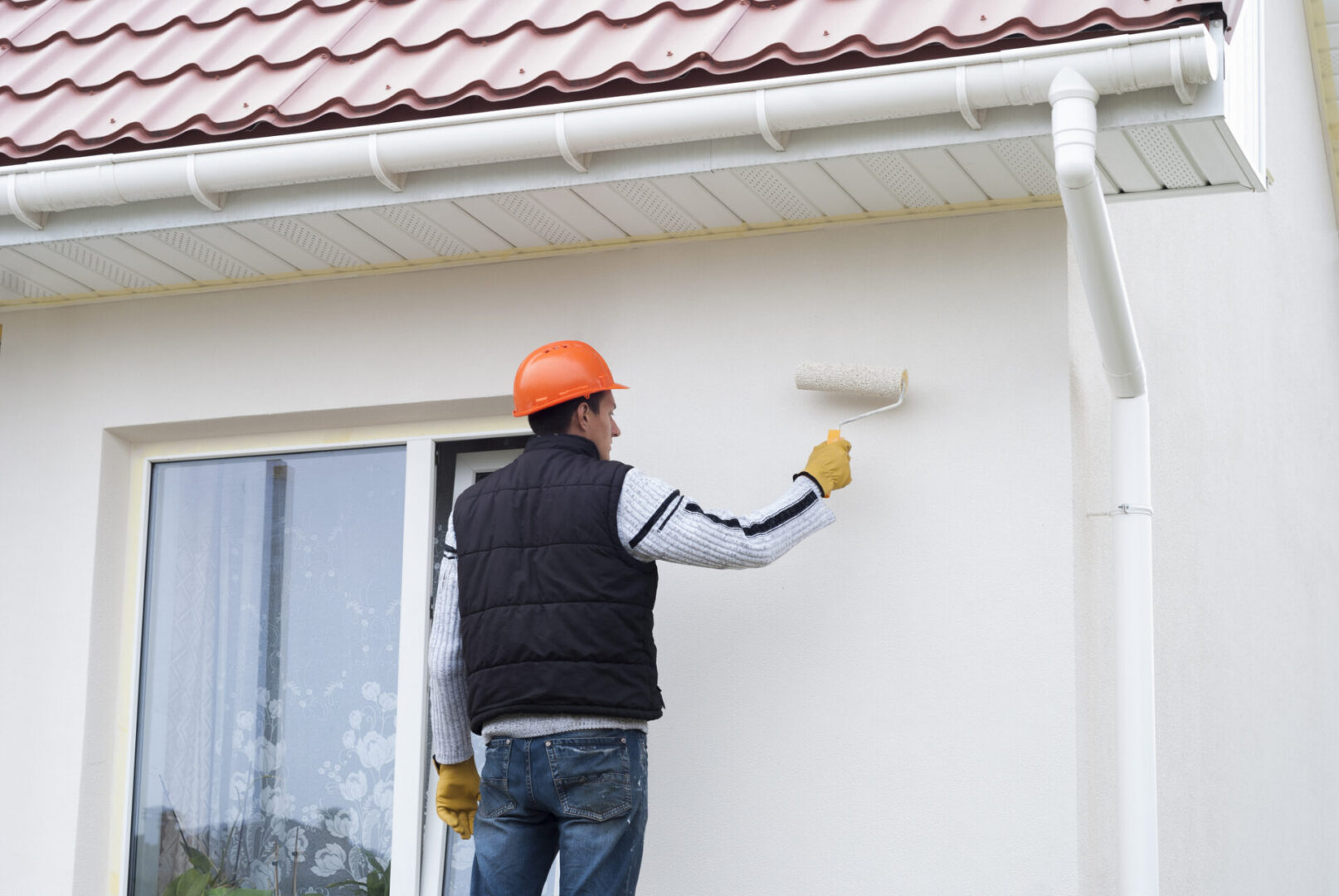 Man wearing a helmet and gloves, painting the side of a house with a paint roller
