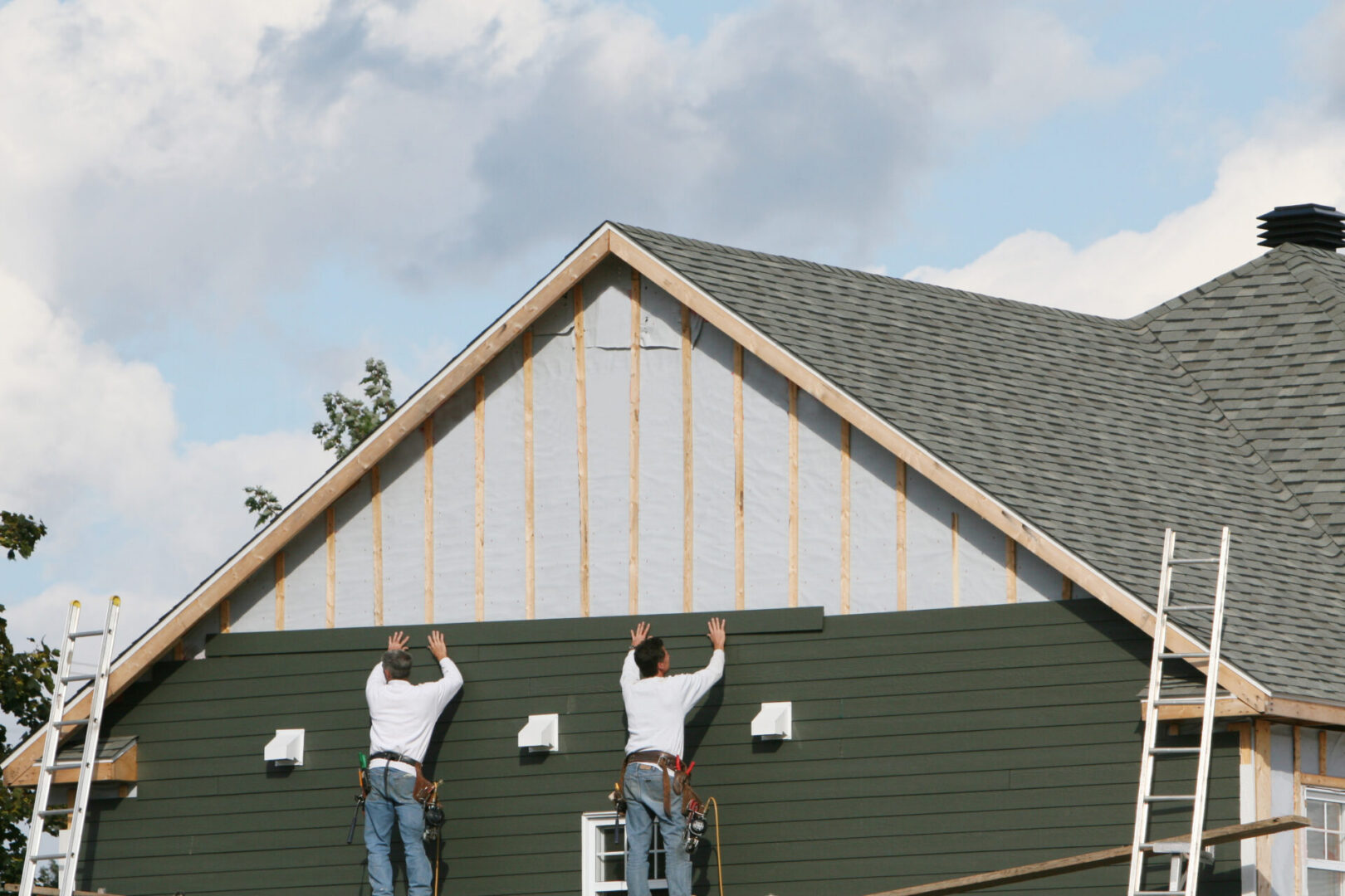 Two men standing on ladders on the roof of a house, surrounded by trees and a clear blue sky