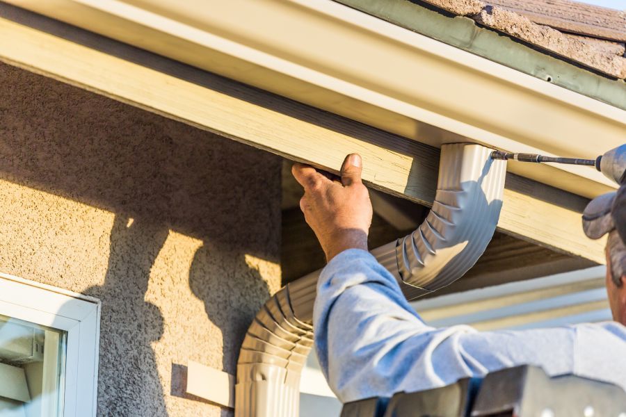 Man wearing a cap and holding a drilling machine, working on the gutter of a house with a window