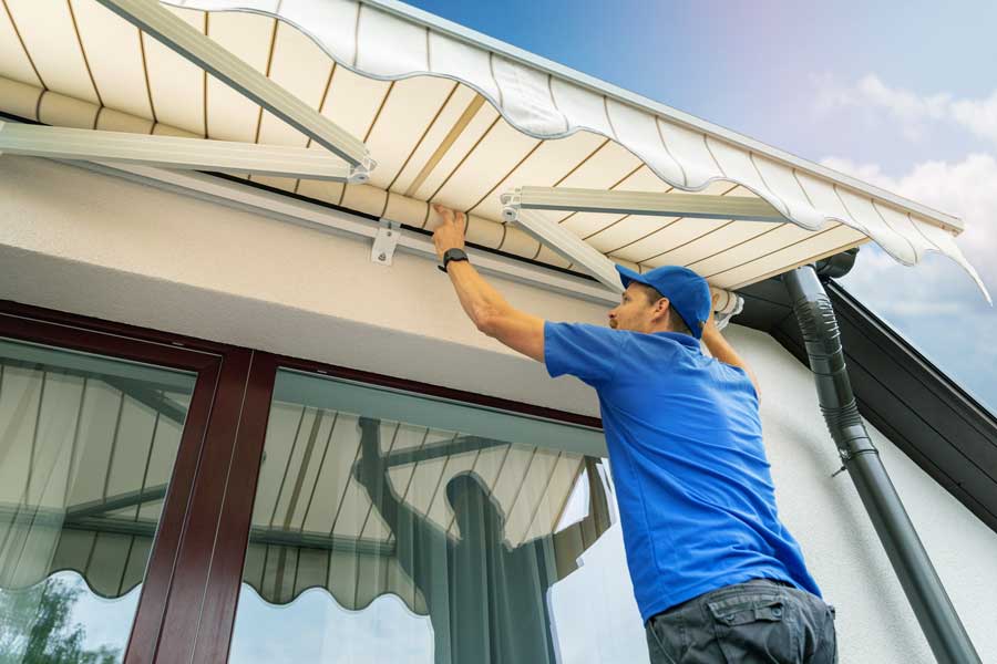 Man wearing a t-shirt, pants, and a cap, installing a retractable awning on the side of a house
