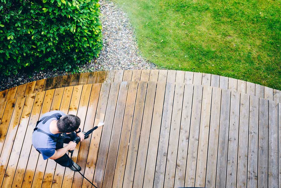 Man using a pressure washer to clean a wooden deck surrounded by lush green grass and plants