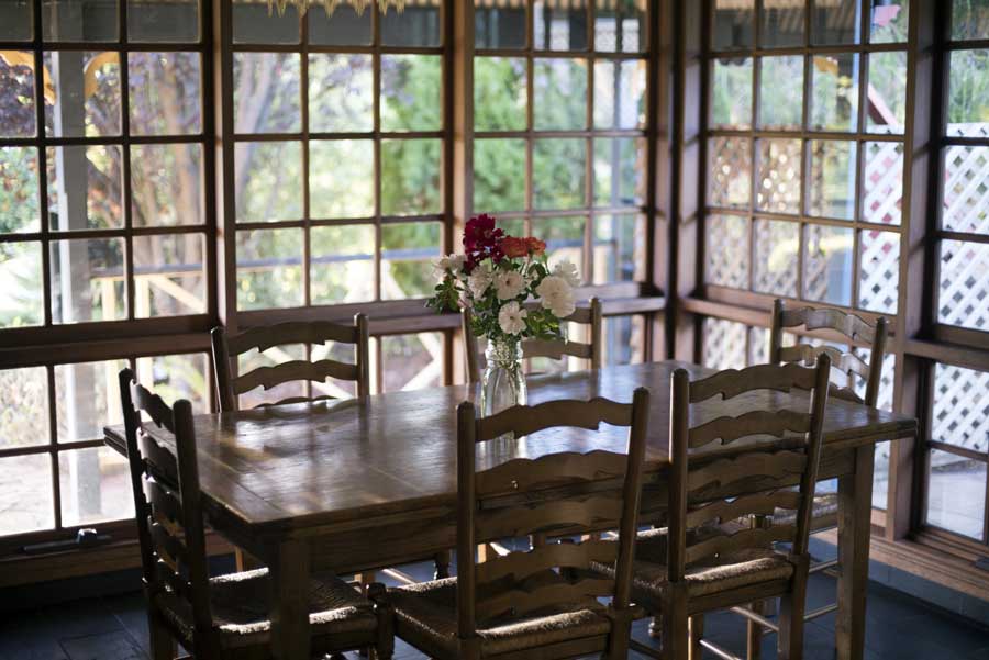 Dining room with a wooden table and chairs, a flower vase on the table, and glass windows in the background