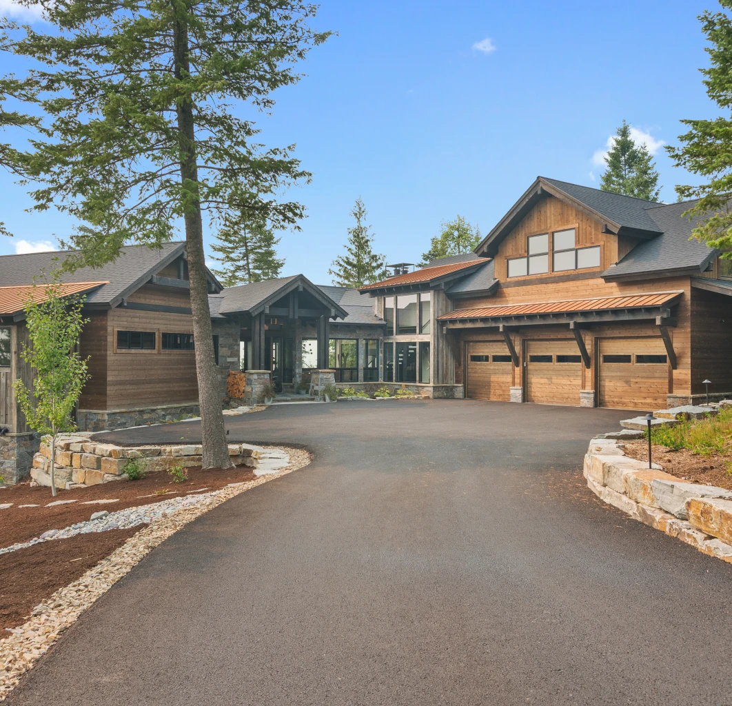 Large house with a driveway in front of it, surrounded by trees, plants, and stones