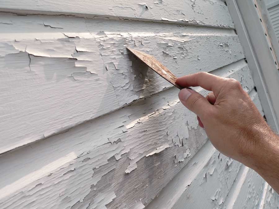 Person using a knife to remove paint from a house siding, likely to repair the siding