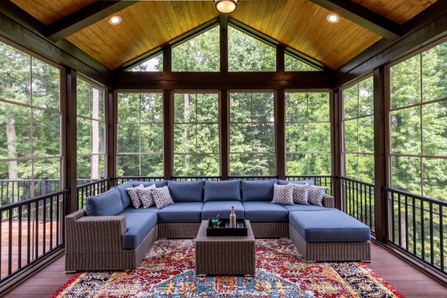 Screened porch with a couch and pillows, a table with bottles and other objects, a carpet on the floor, railings