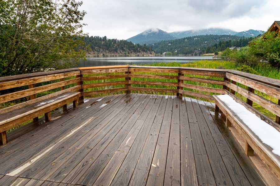 Wooden deck with benches overlooking a lake surrounded by trees, plants, and grass, with a house in the background