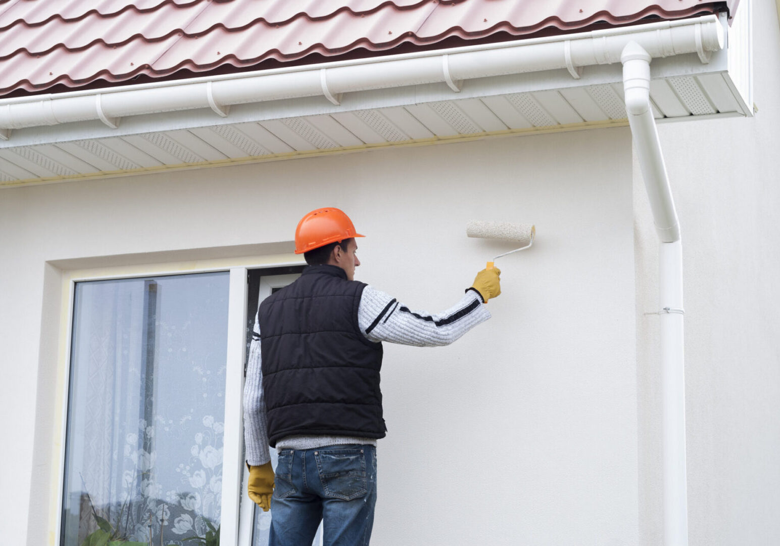 Man wearing a helmet and gloves, painting the side of a house with a paint roller