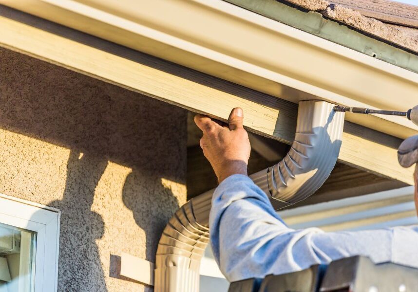 Man wearing a cap and holding a drilling machine, working on the gutter of a house with a window