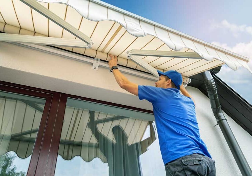 Man wearing a t-shirt, pants, and a cap, installing a retractable awning on the side of a house