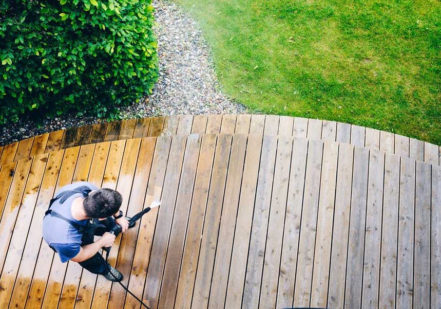 Man using a pressure washer to clean a wooden deck surrounded by lush green grass and plants