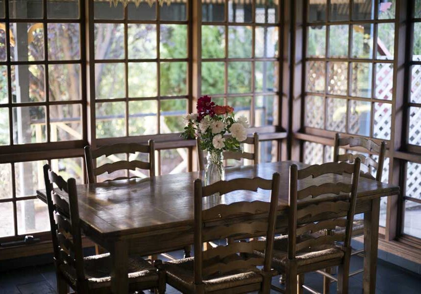 Dining room with a wooden table and chairs, a flower vase on the table, and glass windows in the background