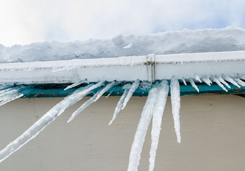 Icicles hanging from the roof of a house, with snow in the background and a clear sky above