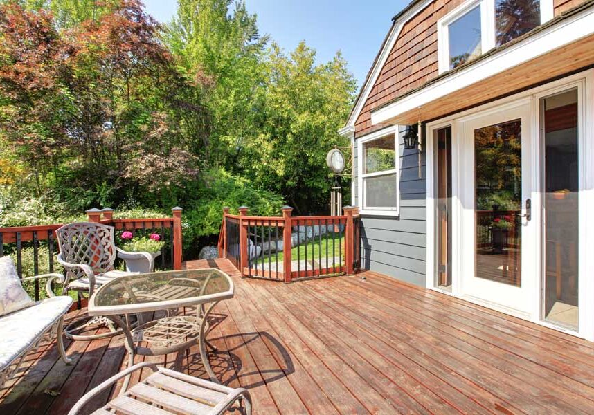 House with glass doors and windows, a wooden deck with a table and chairs on it, a sofa with pillows