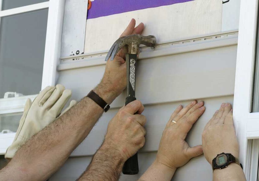 Two men working on a window with a hammer, installing siding