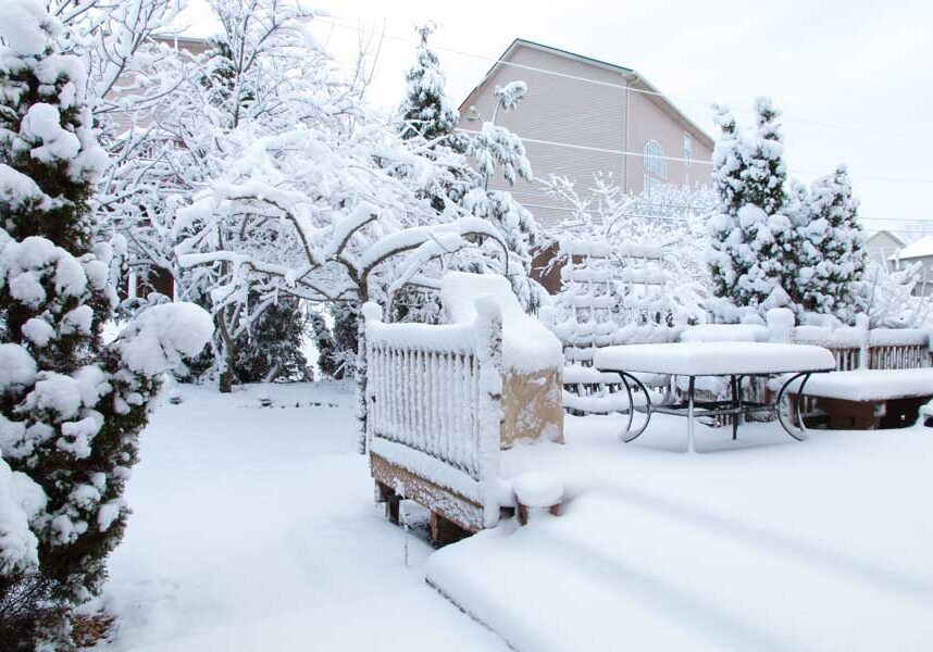 Snow covered patio with a table and chairs surrounded by trees and buildings in the background