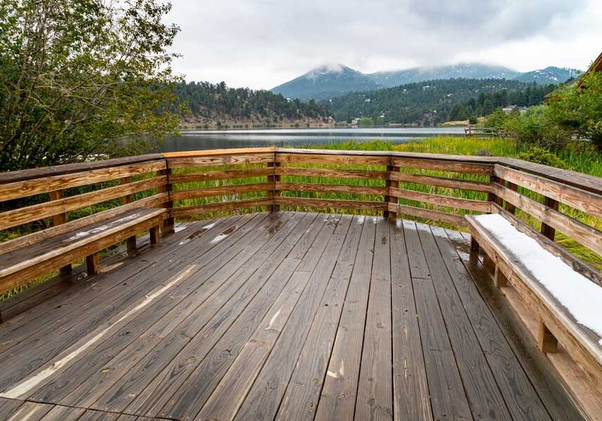 Wooden deck with benches overlooking a lake surrounded by trees, plants, and grass, with a house in the background